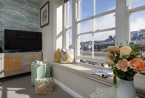 A room with a television on a cabinet, a window with a view of a town, and a vase with flowers on the windowsill. - Five - La Petite Maison is a stylish waterfront holiday apartment in Brixham (Brixham)