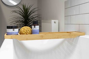 Ein Badezimmer mit einem hölzernen Tablett, auf dem ein Körperbalsam, ein Schwamm und eine Flasche Badesalz liegen.