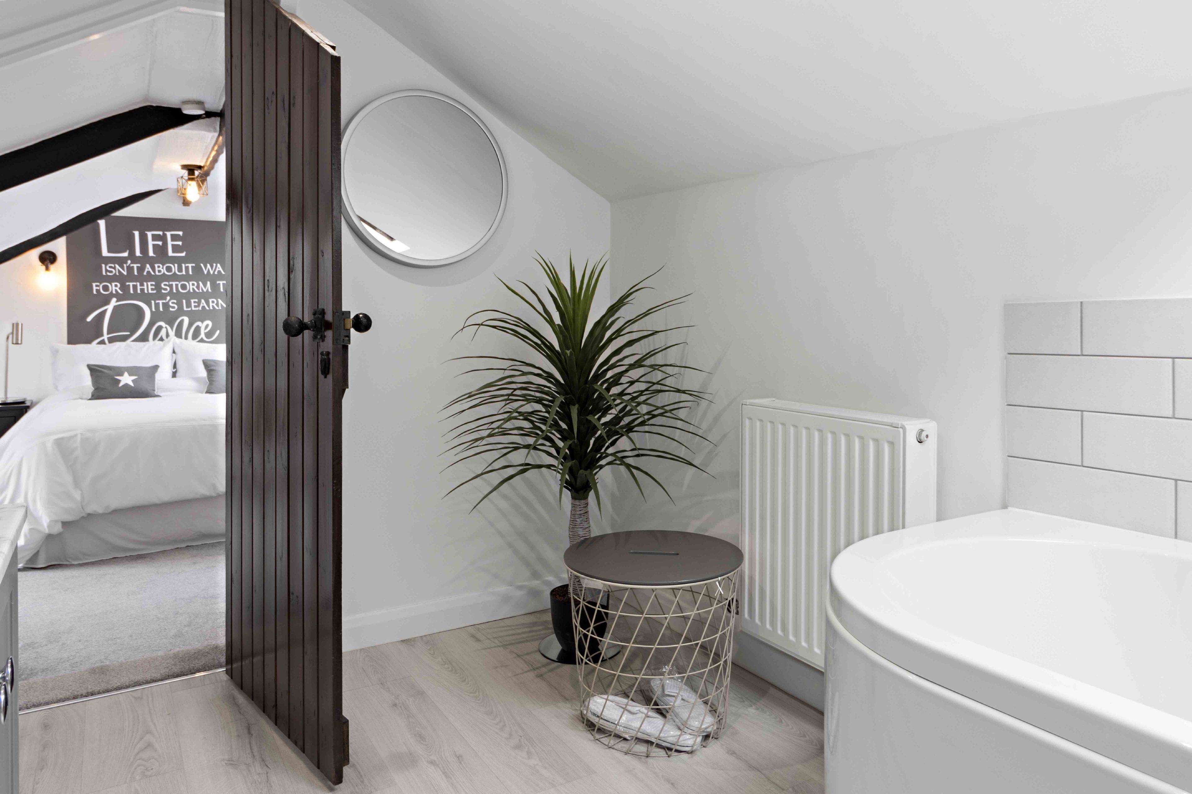 A modern bathroom with a white bathtub, a round mirror, a potted plant, and a motivational quote on the wall.
