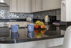 A kitchen with a black countertop, a fruit basket, and two blue mugs with white letters. - Bayview Retreat is a stunning one bedroom sea view apartment in Torquay (Torquay)