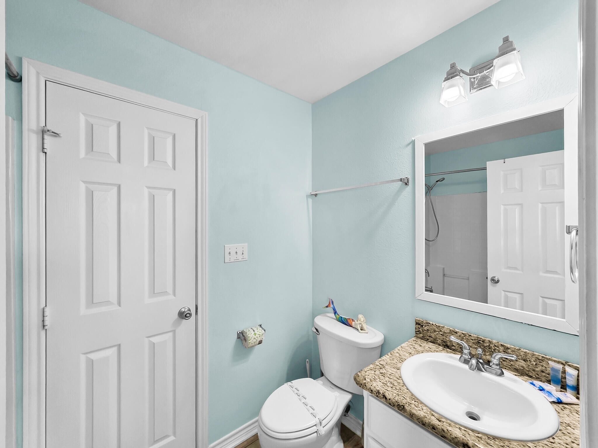 A bathroom with a white door, a toilet, a sink with a granite countertop, and a mirror above the sink.
