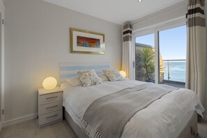 Ein Schlafzimmer mit einem Bett, Nachttischlampen, einem Nachttisch, einem gerahmten Bild und Meerblick.