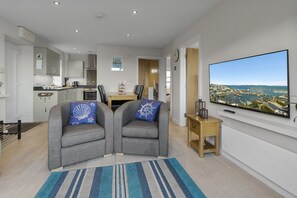 Ein modernes Wohnzimmer mit Fernseher, zwei grauen Sesseln mit marineblauen Kissen, einem Holztisch und Blick auf eine Küstenstadt.