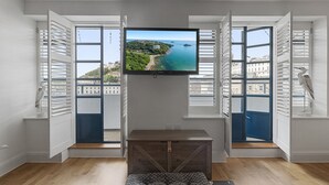 A room with a TV mounted on a wall, a wooden bench, a pet bed, and a window with a view of a coastal area. - Harbour Lights Apartment, a coastal retreat set on Torquay's vibrant marina (Torquay)