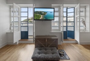 A room with a TV mounted on a wall, a wooden bench, a pet bed, and a window with a view of a coastal area. - Harbour Lights Apartment, a coastal retreat set on Torquay's vibrant marina (Torquay)