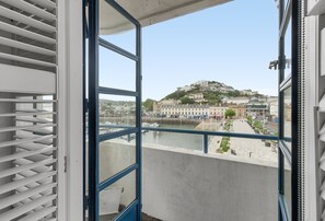 Blick aus einem Fenster mit Jalousien auf einen Wasserblick mit Gebäuden und einem Hügel.
