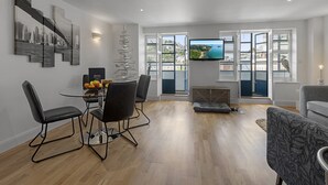 A modern living room with a dining area, a TV mounted on the wall, and a view of a coastal landscape through large windows. - Harbour Lights Apartment, a coastal retreat set on Torquay's vibrant marina (Torquay)