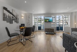 A modern living room with a dining area, a TV mounted on the wall, and a view of a coastal landscape through large windows. - Harbour Lights Apartment, a coastal retreat set on Torquay's vibrant marina (Torquay)