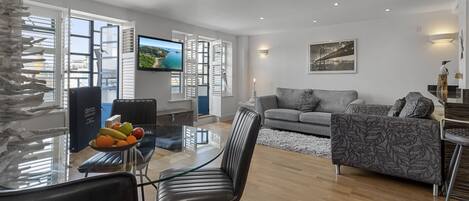 A modern living room with a glass dining table, a grey sofa, and a flat-screen TV.