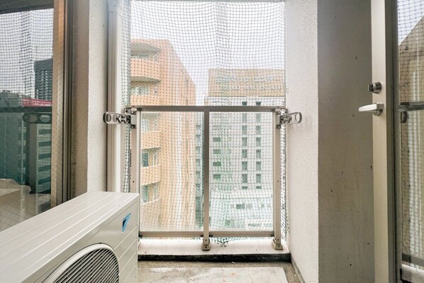 A balcony with a metal railing, a view of a cityscape, and a white air conditioning unit. - Trioni EBISU (Osaka)