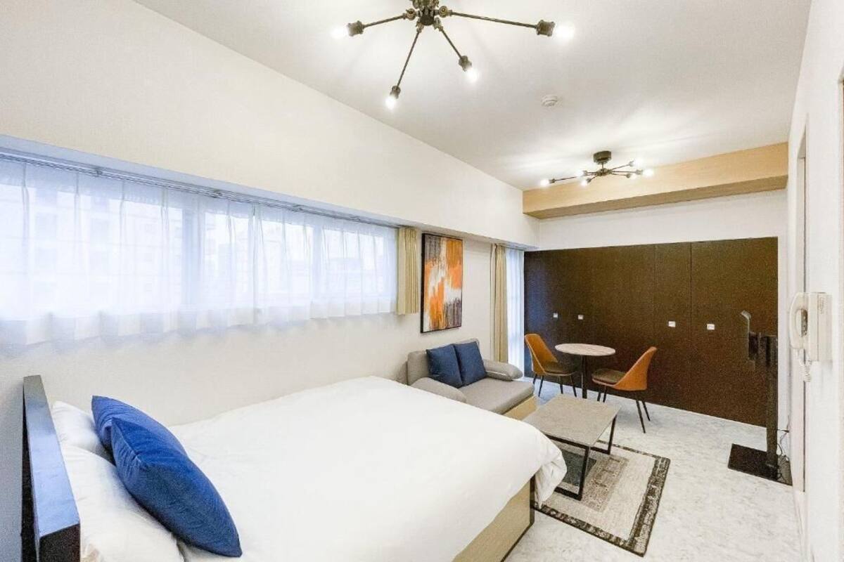 Une chambre d’hôtel avec un lit, un canapé, une petite table et une chaise. Il y a une fenêtre avec des rideaux, un tableau au mur et un luminaire au plafond.
