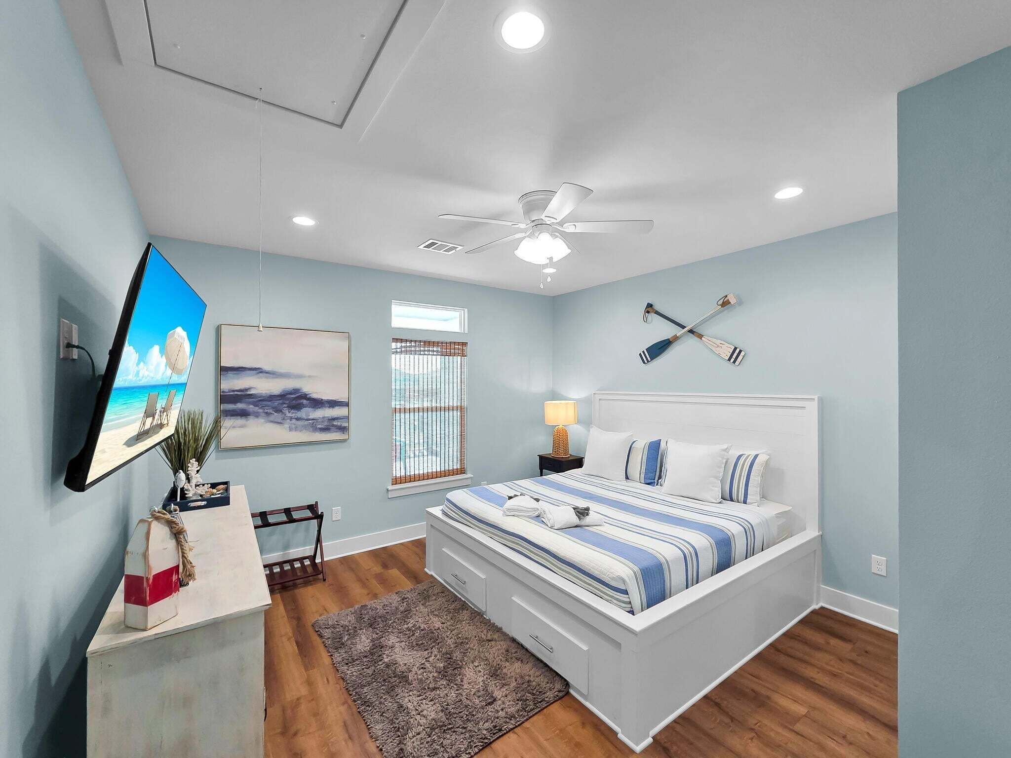 Un dormitorio con cama, ventilador de techo, televisor, ventana con persianas e imagen colgada en la pared.