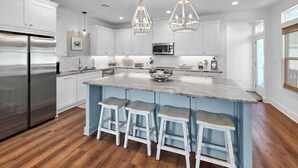 A modern kitchen with white cabinets, a stainless steel refrigerator, a large island with bar stools, pendant lighting, and a microwave. - Pearly Pelican 3 Bedroom Home (Port Bolivar)