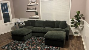 A sectional sofa, a patterned rug, a small table, and a potted plant in a room with wooden flooring and a window.