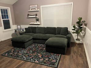 A sectional sofa, a patterned rug, a small table, and a potted plant in a room with wooden flooring and a window. - 3-bedroom house in gorgeous Lawrence with easy access to K-10 and I-70 (Lawrence)