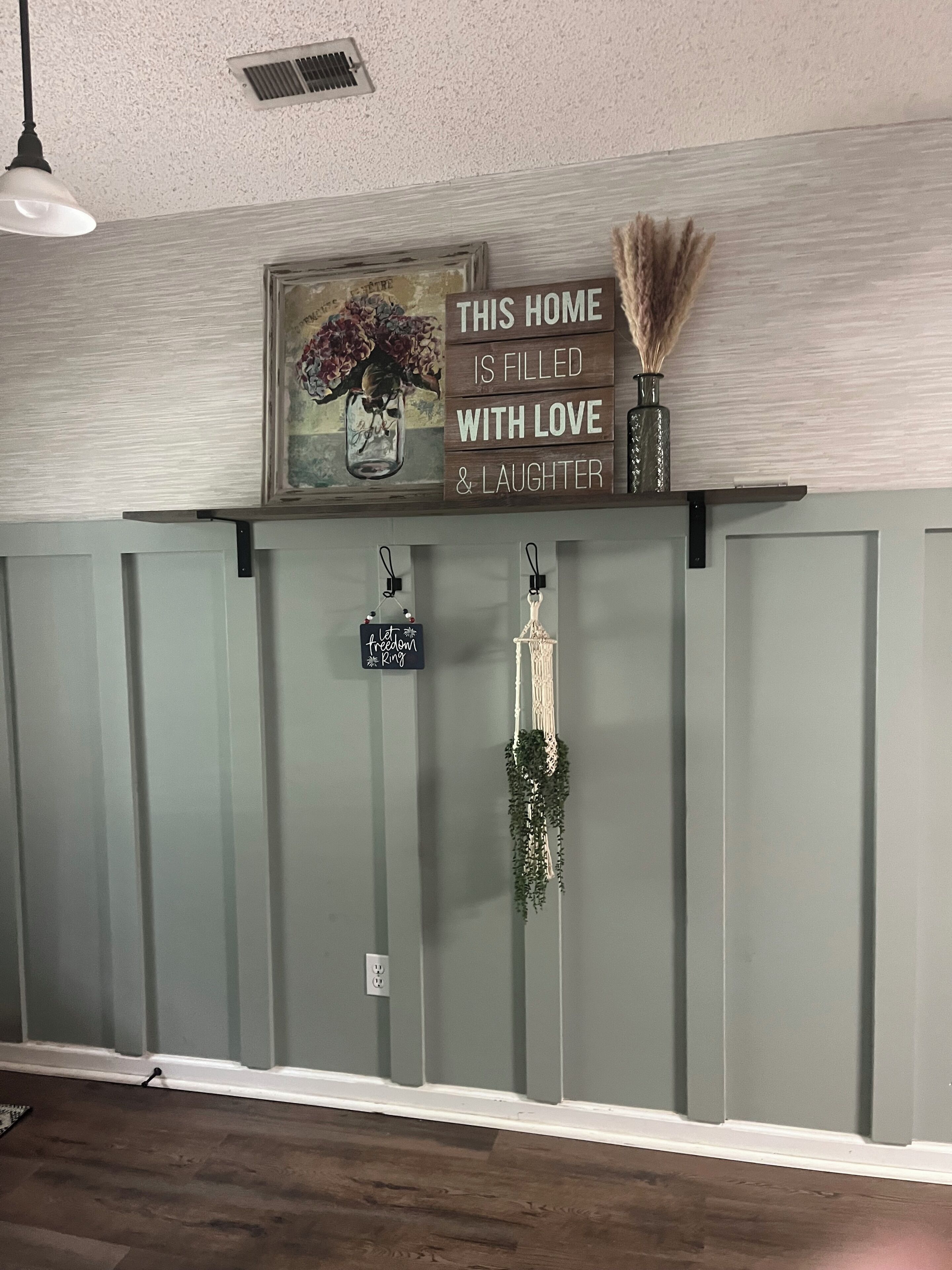 Un mur orné d’une pancarte indiquant « CETTE MAISON EST REMPLIE D’AMOUR ET DE JOIE », d’un tableau encadré et de deux décorations en macramé.