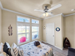 A bedroom with a bed, a ceiling fan, a window with a view, a door, and a small shelf. - It's A Shellabration 3 Bedroom Home (Port Bolivar)