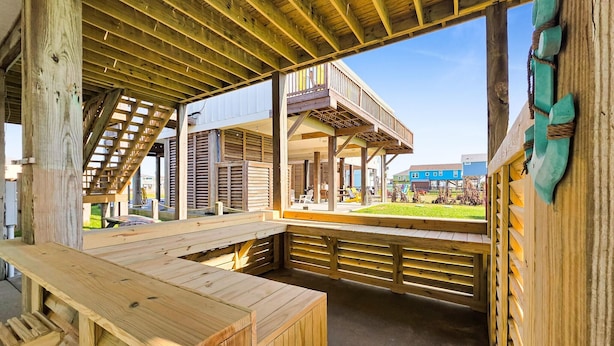A wooden deck with a bench, a covered area with a view of a blue building, and a wooden structure with a staircase. - It's A Shellabration 3 Bedroom Home (Port Bolivar)
