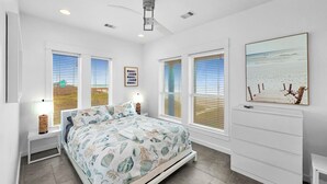 A bedroom with a bed, a ceiling fan, a nightstand, a lamp, and a painting on the wall. - Crystal By The Ocean 4 Bedroom Home (Port Bolivar)