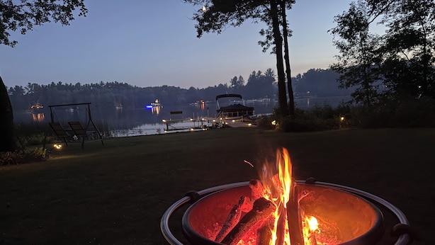 Ein Lagerfeuer in einer Metall-Feuerstelle mit einem See und Gebäuden im Hintergrund.