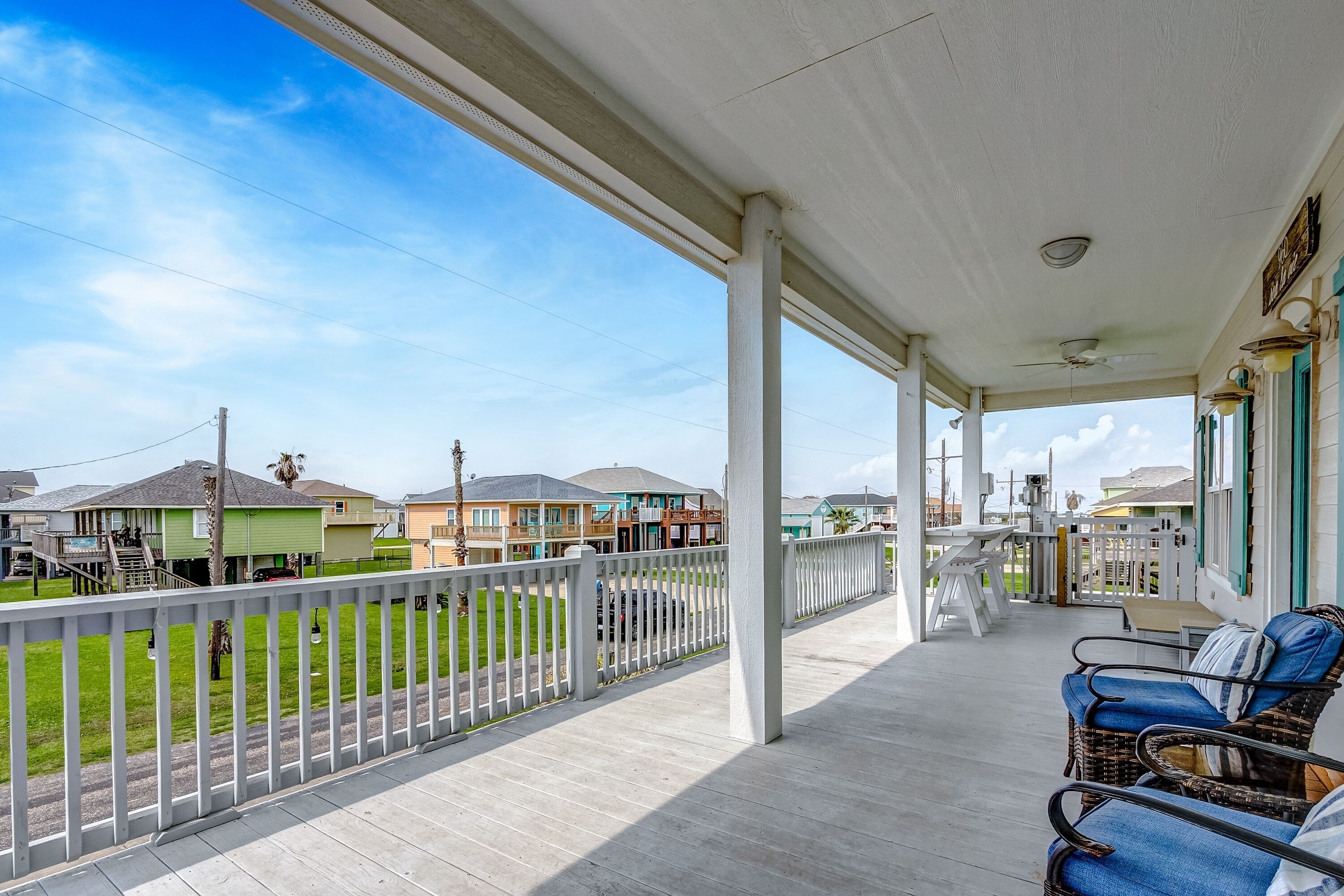 Een overdekt terras met een witte railing, rieten meubels en uitzicht op omliggende huizen.