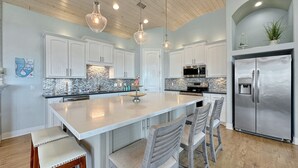 A modern kitchen with white cabinets, a stainless steel refrigerator, and a central island with bar stools. - The Salty Mermaid 3 Bedroom Home (Port Bolivar)