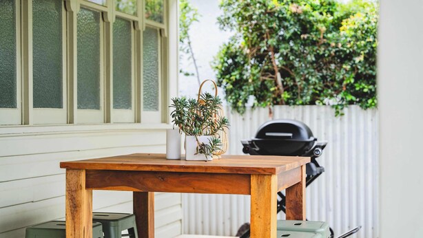 Een houten tafel met een bloempot, twee groene krukken en een barbecue op de achtergrond.