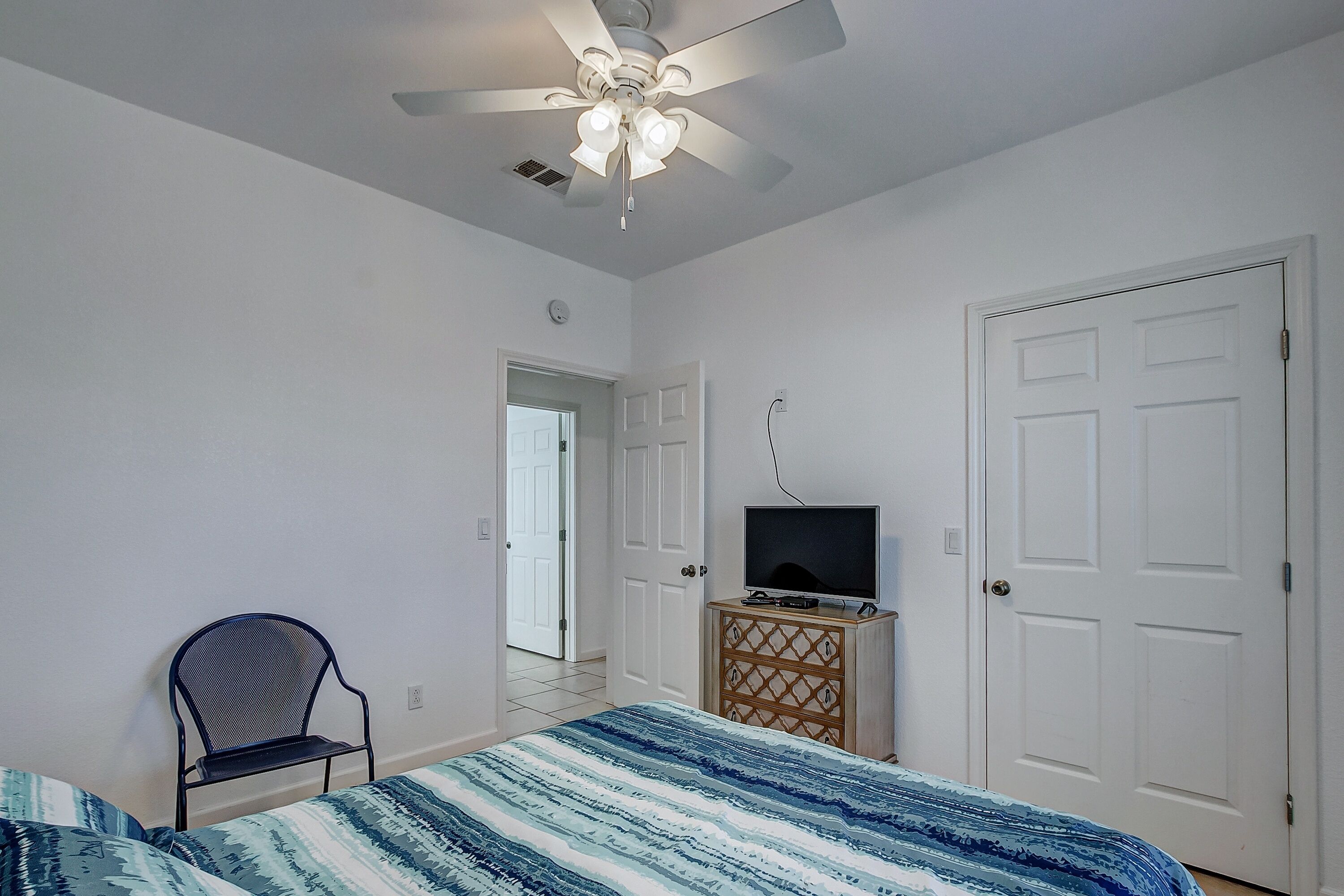 A bedroom with a bed, a chair, a TV on a wooden cabinet, a ceiling fan, and white doors.