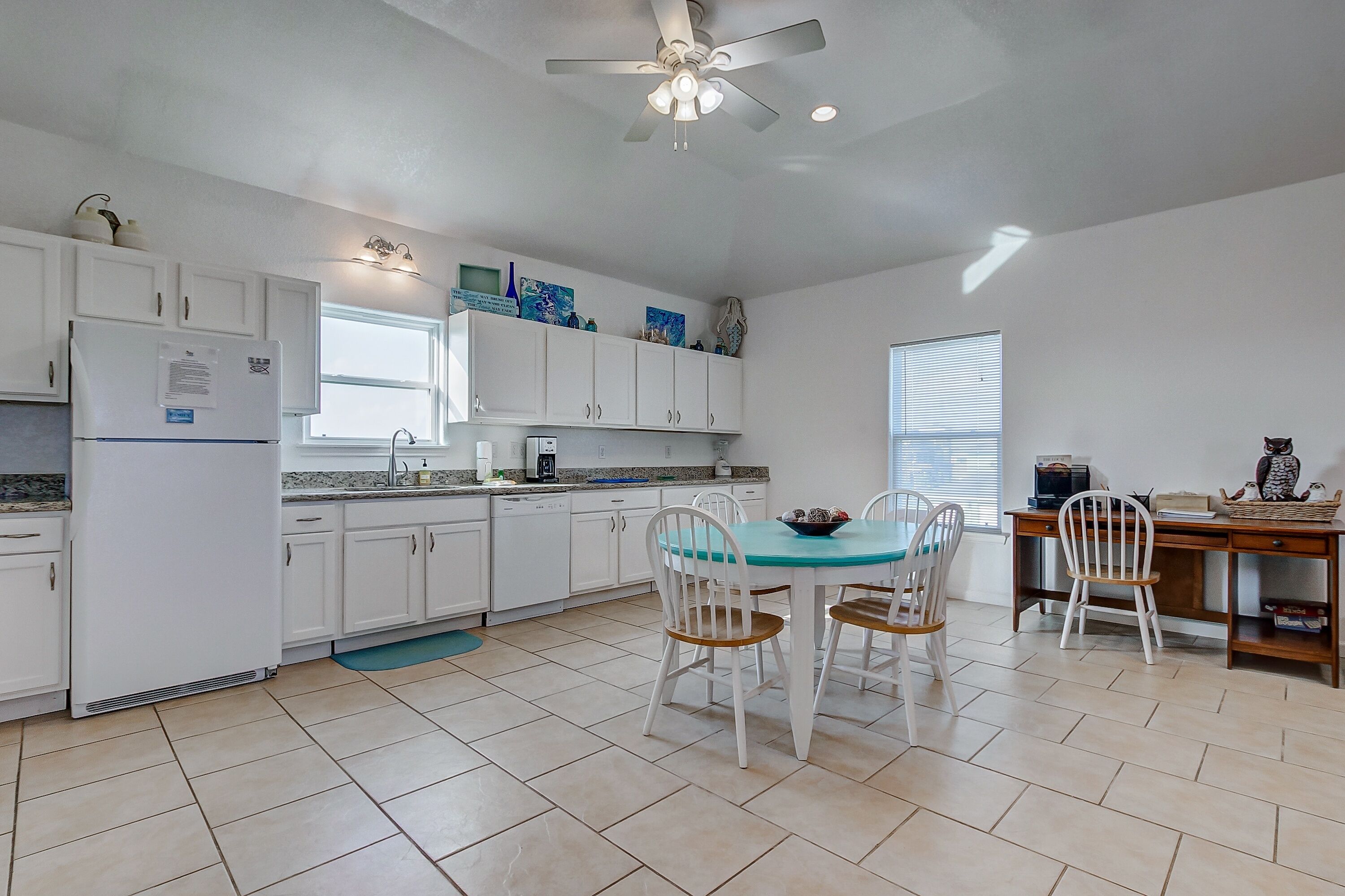 A kitchen with white cabinets, a dining table with chairs, a ceiling fan, and a window.