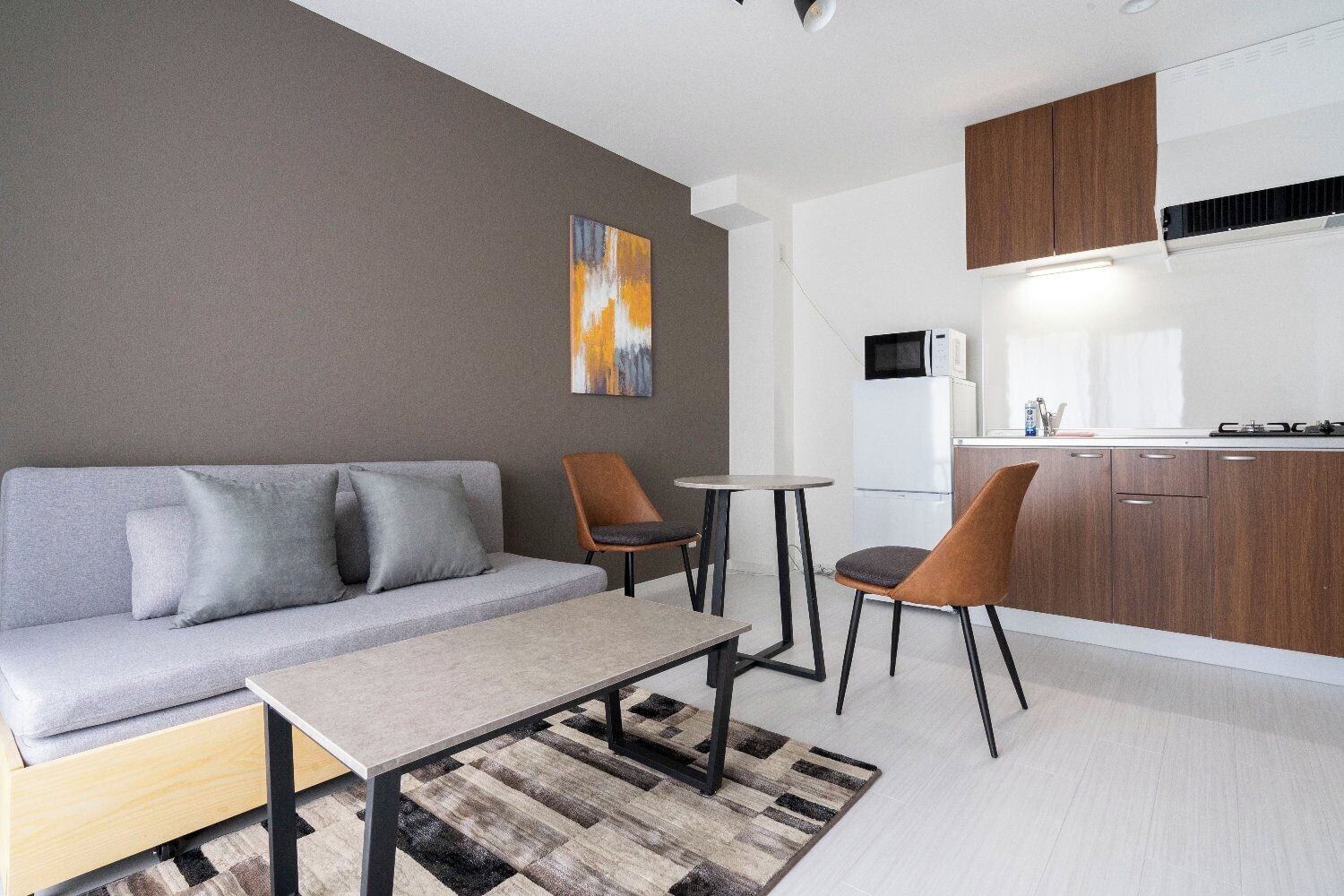 Una sala moderna con un sofá, dos sillones, una mesa de centro, una zona de cocina con armarios y un microondas, y un cuadro en la pared.