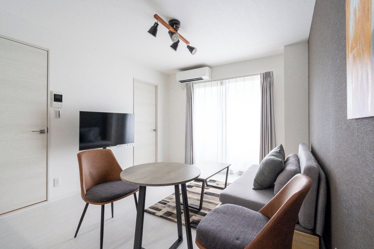 A modern living room with a sofa, two chairs, a round table, a flat-screen TV, and a ceiling light fixture.