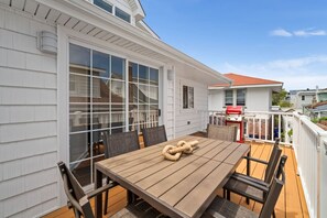 A wooden deck with a table and chairs, a grill, and a white house with large windows. - Spacious 6BR - Sleeps 18| Beach Gear | Deck | BBQ (Ventnor City)