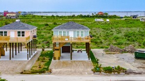 Two waterfront houses with elevated decks, a sandy beach, and a body of water in the background. - A Quietude 3 Bedroom Home (Port Bolivar)