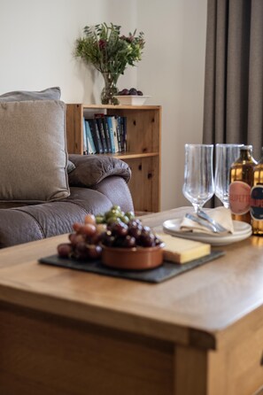 Ein gemütliches Wohnzimmer mit einer Couch, einem Holztisch mit einer Schale Trauben, einem Weinglas und einer Flasche.