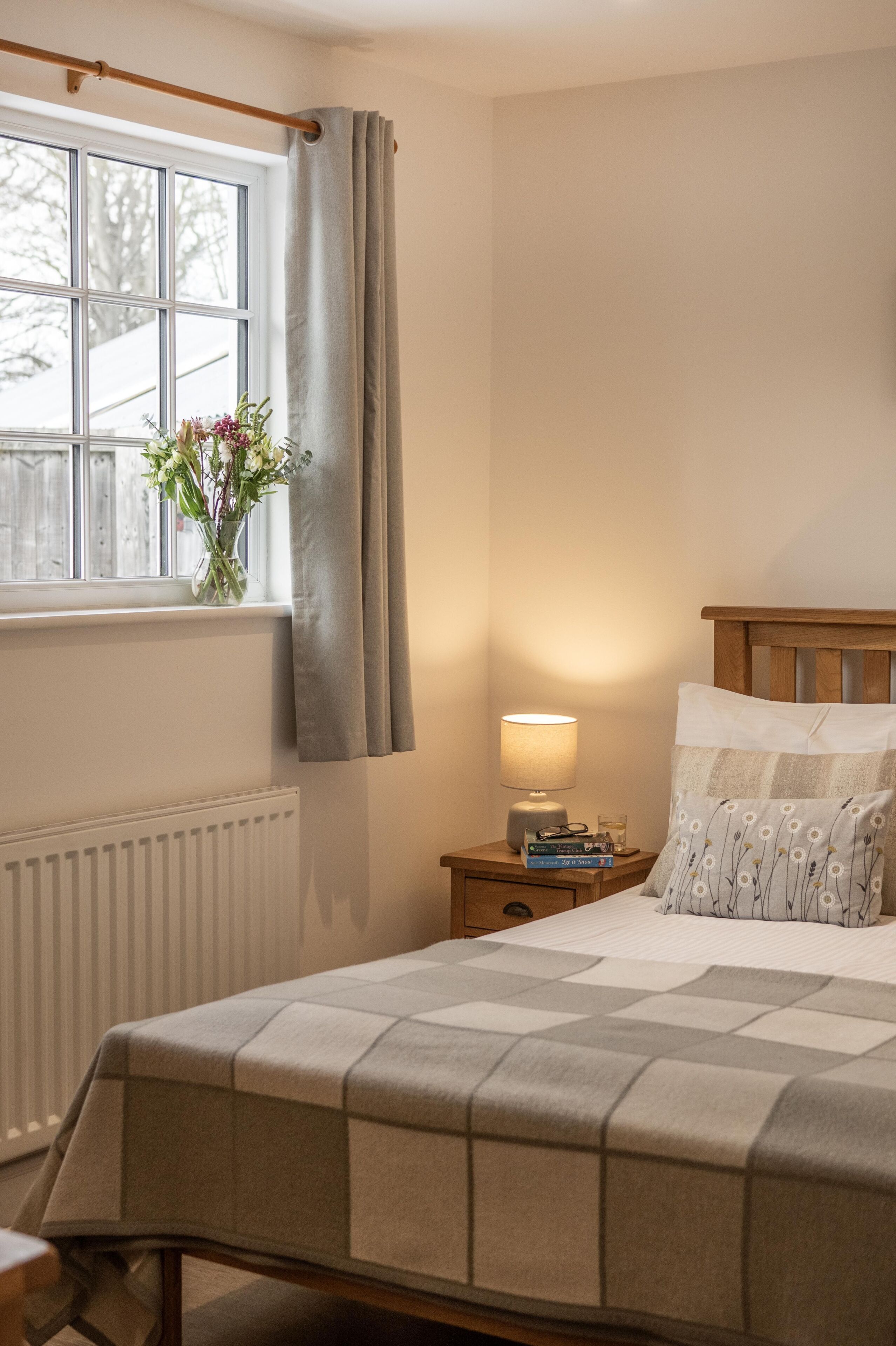 A bedroom with a bed, a nightstand, a lamp, a window with curtains, and a radiator.