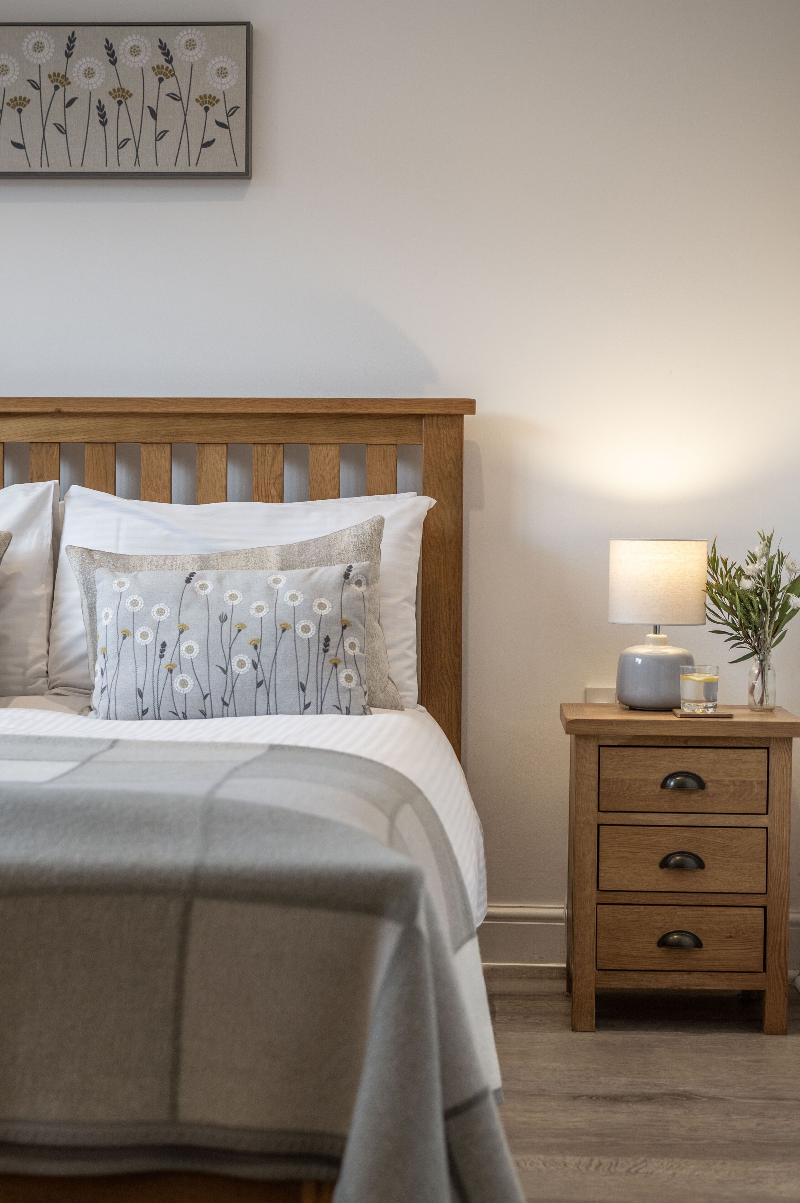A neatly made bed with a floral pillow, a bedside lamp, and a small plant on a wooden nightstand.