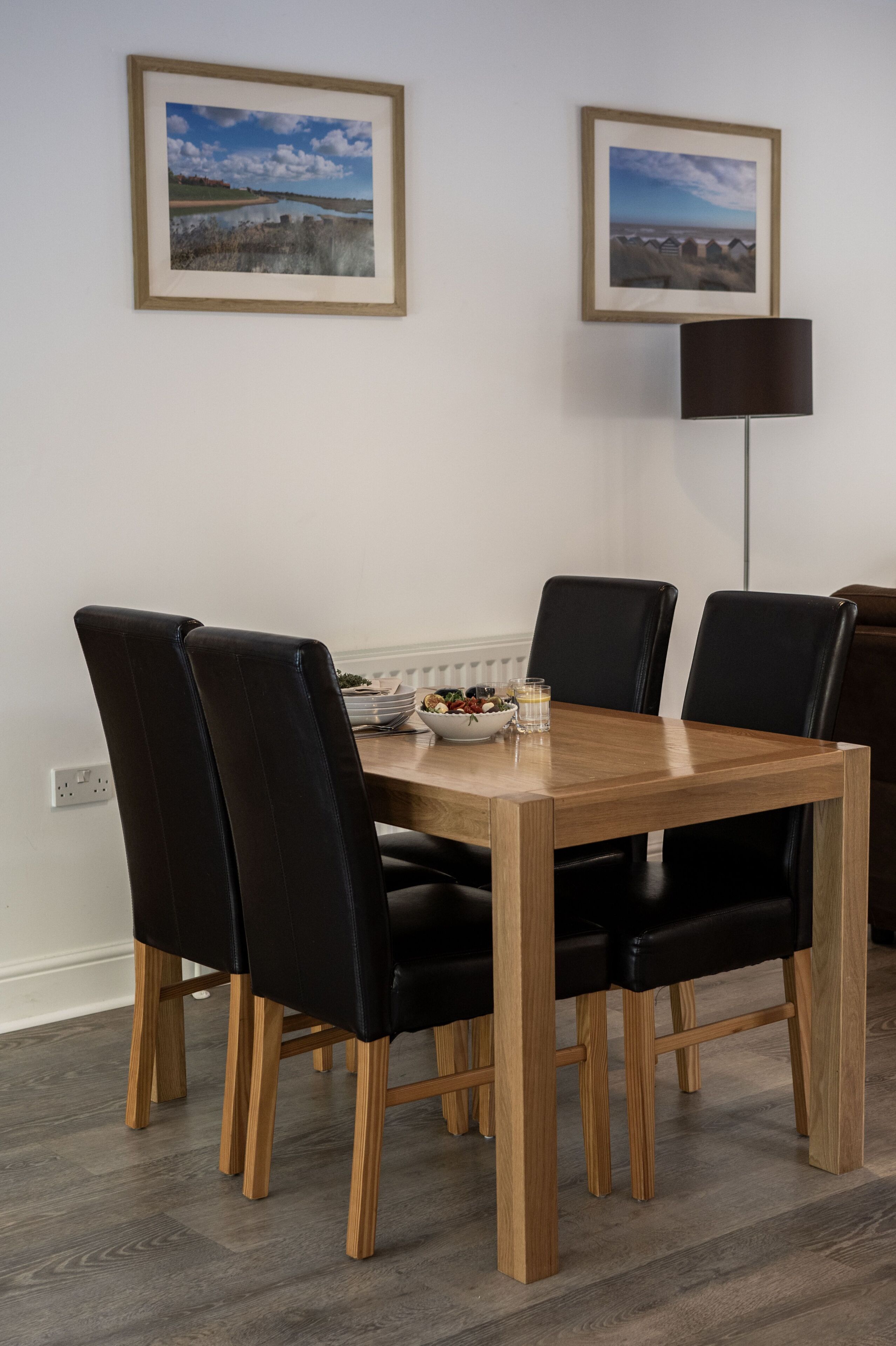 Una mesa de comedor con seis sillas negras, un cuenco de fruta y una vela sobre la mesa. Dos cuadros enmarcados decoran la pared sobre la mesa.