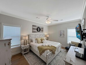 Ein Schlafzimmer mit einem Bett, Nachttischen, einem Deckenventilator, einem an der Wand montierten Fernseher und einem Fenster mit Jalousien.