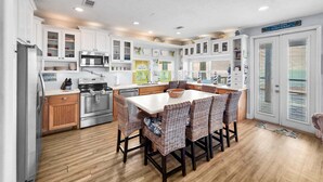 A modern kitchen with white cabinetry, a central island with wicker chairs, stainless steel appliances, and a view of the outdoors through a door. - A Stairway To Heaven 4 Bedroom Home (Port Bolivar)