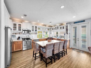 A modern kitchen with white cabinetry, a central island with wicker chairs, stainless steel appliances, and a view of the outdoors through a door. - A Stairway To Heaven 4 Bedroom Home (Port Bolivar)