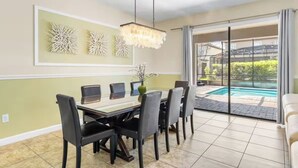 Un coin repas avec une table, des chaises et un lustre. On peut apercevoir une piscine à travers les portes vitrées.