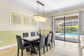 A dining area with a table, chairs, and a chandelier. There is a pool visible through the glass doors. - 9BR Luxury Disney Villa w/ Pool, Game Room & Resort Access (Four Corners)