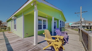A green building with a porch, two Adirondack chairs, and a wooden deck. - Lime In The Coconut 3 Bedroom Home (Port Bolivar)