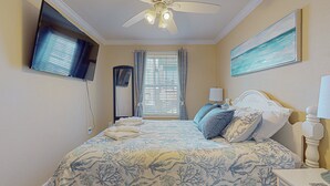 Habitación de hotel con cama, televisión montada en la pared, ventilador de techo, ventana con cortinas y un cuadro en la pared.