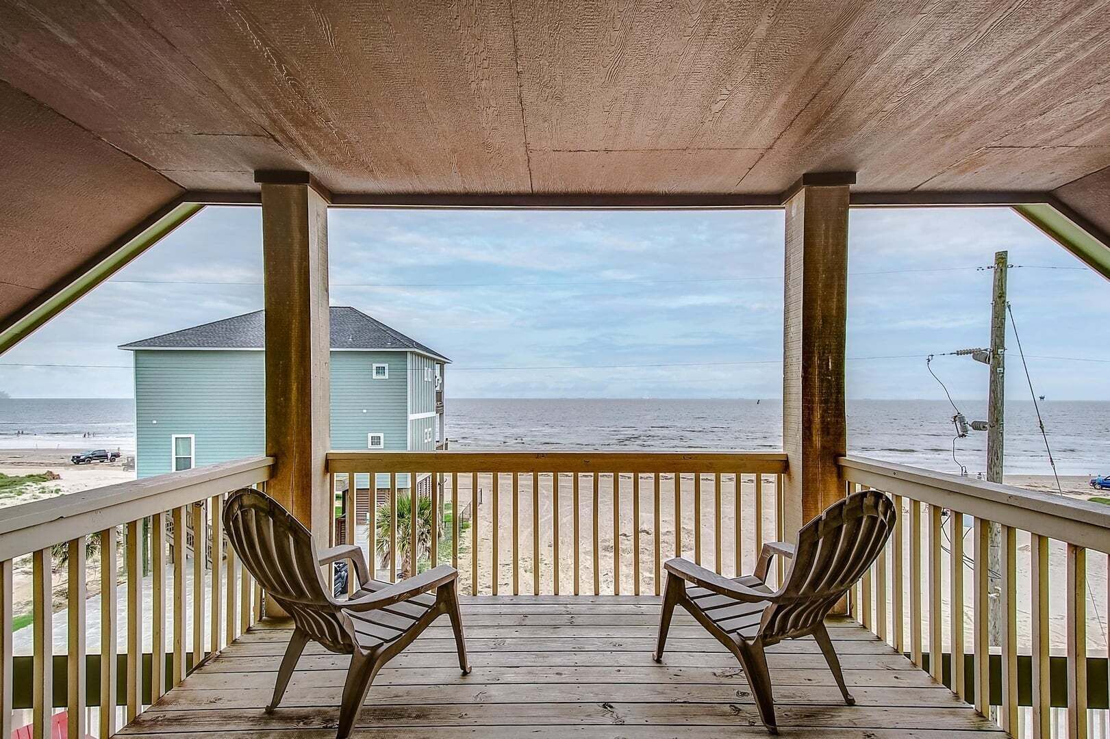 A covered porch with two chairs, a view of the beach, and a house in the background.