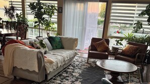 A spacious living room with a large window, a sofa, a coffee table, and various plants.