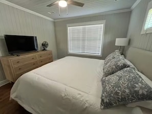 Ein Schlafzimmer mit Bett, Kommode, Fernseher, einem Fenster mit Jalousien und einem Deckenventilator.
