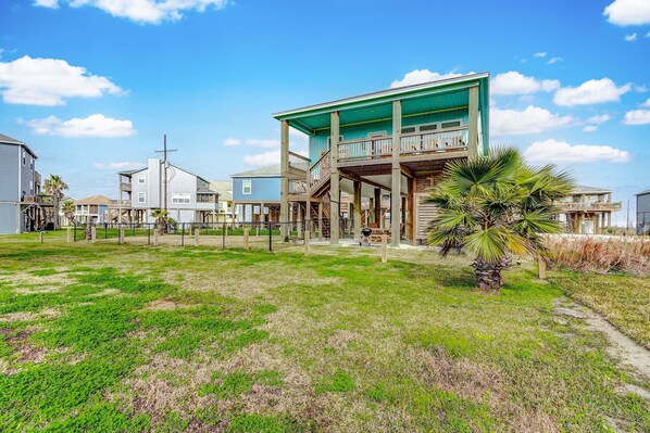 A beachfront property with a large house featuring a balcony, a covered porch, and a small garden area with palm trees. - Afterdunedelight 3 Bedroom Home (Port Bolivar)