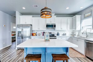 A modern kitchen with white cabinets, stainless steel appliances, a central island with bar stools, and a unique pendant light. - Afterdunedelight 3 Bedroom Home (Port Bolivar)