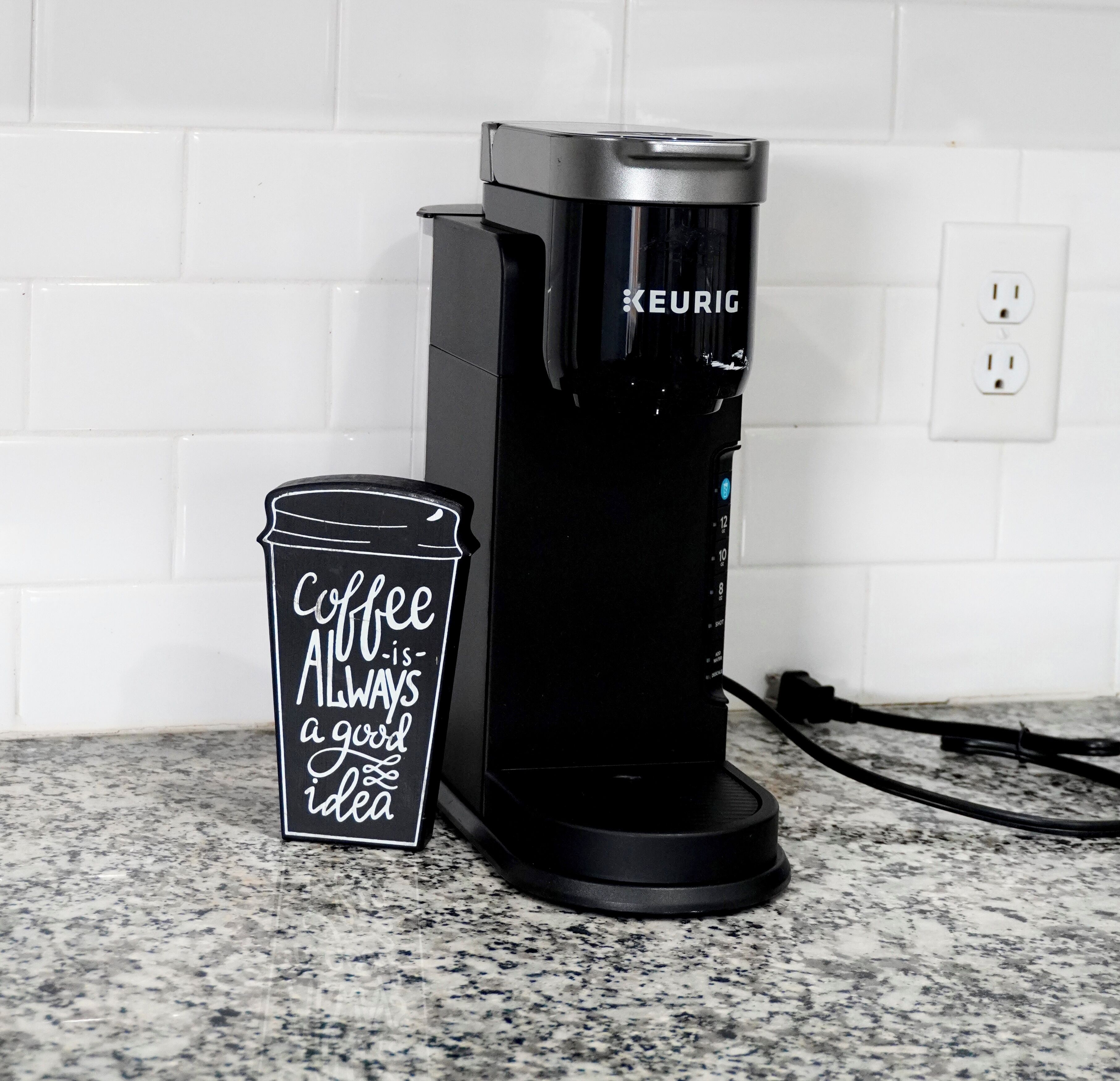 Une machine à café Keurig posée sur un plan de travail de cuisine, à côté d’un gobelet jetable sur lequel on peut lire : « Le café, c’est toujours une bonne idée. »
